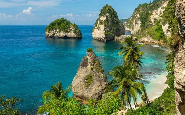 Beautiful coastal view of Nusa Penida with iconic cliffs, rock formations, clear blue ocean, and palm trees.
