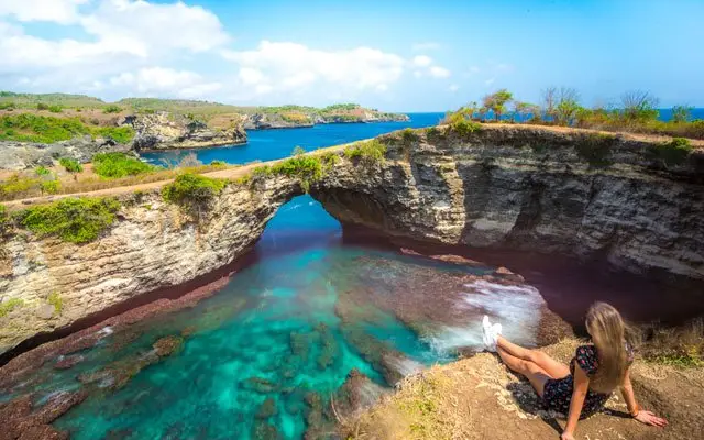 Traveler relaxing near the famous natural rock bridge at Nusa Lembongan, with clear blue water and scenic cliffs, during a trip from Bali to Nusa Lembongan.