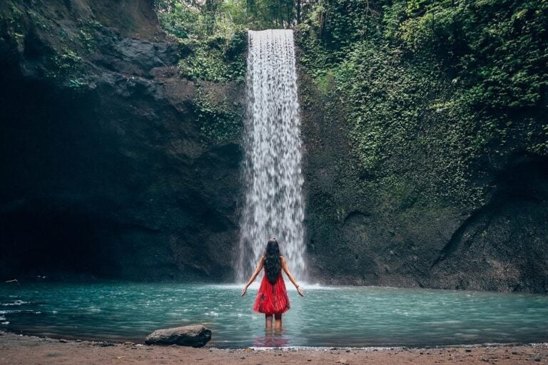 Woman in red dress standing in front of Tibumana Waterfall in Bali, surrounded by lush green forest and clear water pool.